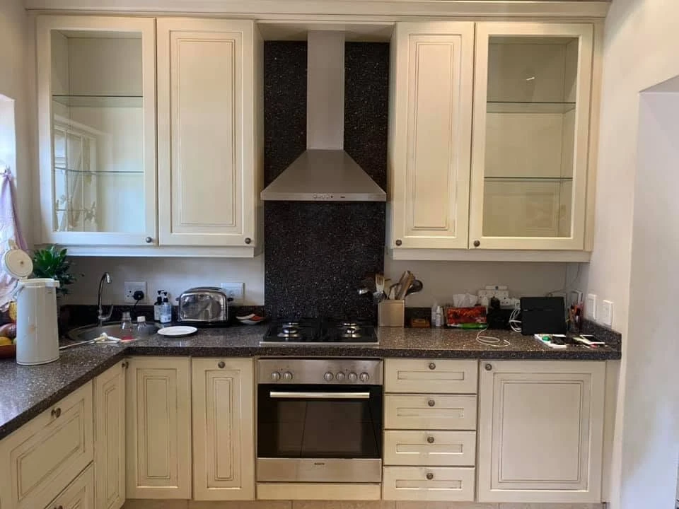 Before — Cream framed cabinetry taken to a deep satin black, with brushed gold handles and warm cabinet lighting. The speckled granite tops, extractor and glass display uppers stay — the same kitchen, an entirely new mood.