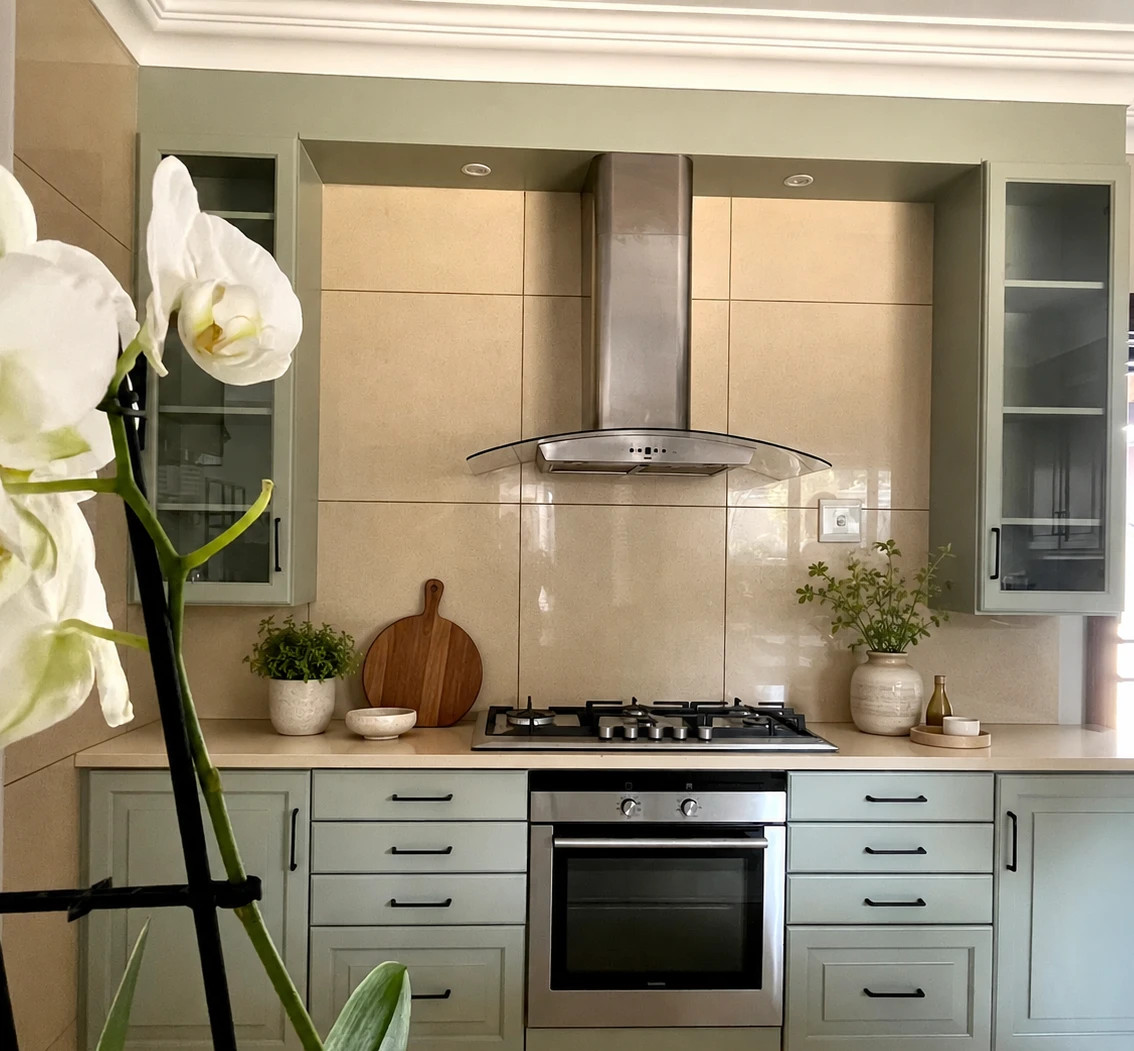 Detail — Honey-toned timber cabinetry hand painted in a soft sage green. Matte black handles, warm under-cabinet lighting and open display shelving bring a calm, country-modern feel — original layout and stone tops retained.