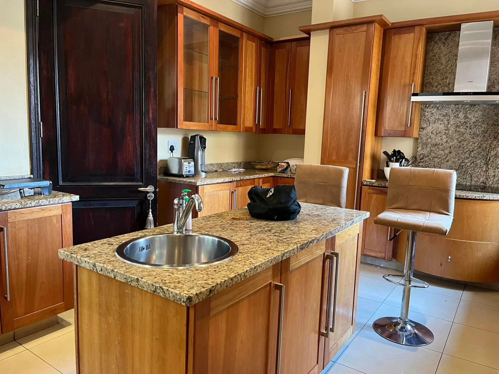 Before — Honey-toned cabinetry refreshed in soft warm beige, with a charcoal island, solid timber top and fluted detail. A black granite sink, matte spring tap and brushed aluminium kickplates complete the look — original layout intact.